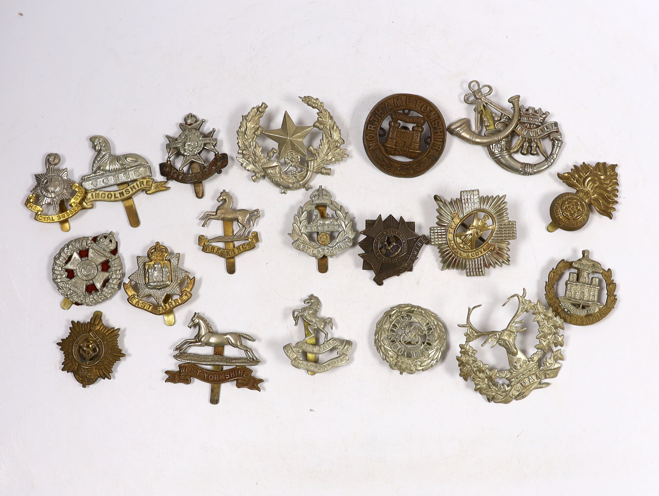 Twenty military cap badges including Bedfordshire, West Yorkshire, Cornwall, East Lancashire, the Royal Scots, the Royal Sussex, Northamptonshire, Lincolnshire, East Surrey, Dorsetshire, Engineer Volunteers, etc.